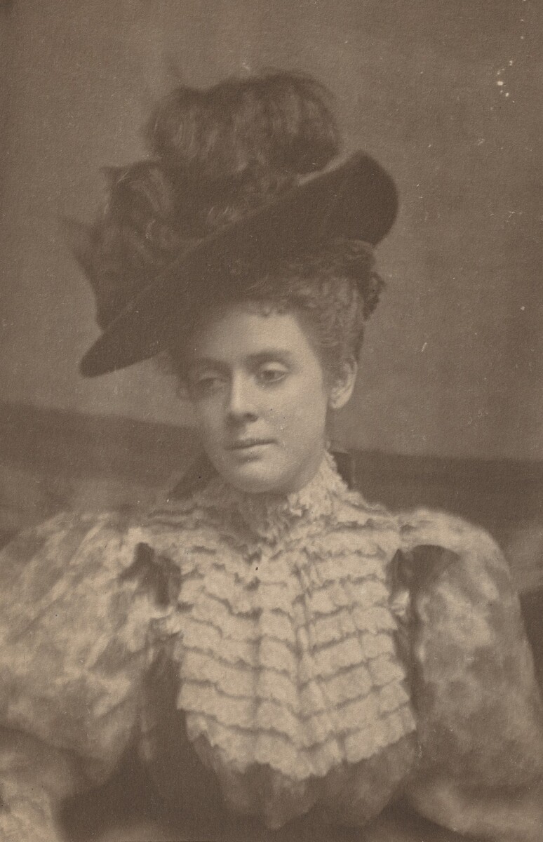 The image depicts a woman seated or slightly leaning, captured from the waist up. Her head is tilted to one side, with her gaze averted from the camera. She has delicate facial features and a serene expression. Her hair is styled in an intricate updo under a large, dark, slightly tilted hat. She wears a high-necked dress with ruffled fabric around the chest and shoulders. The background is plain with a soft-focus texture.
