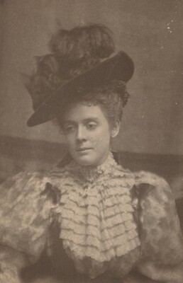 The image depicts a woman seated or slightly leaning, captured from the waist up. Her head is tilted to one side, with her gaze averted from the camera. She has delicate facial features and a serene expression. Her hair is styled in an intricate updo under a large, dark, slightly tilted hat. She wears a high-necked dress with ruffled fabric around the chest and shoulders. The background is plain with a soft-focus texture.