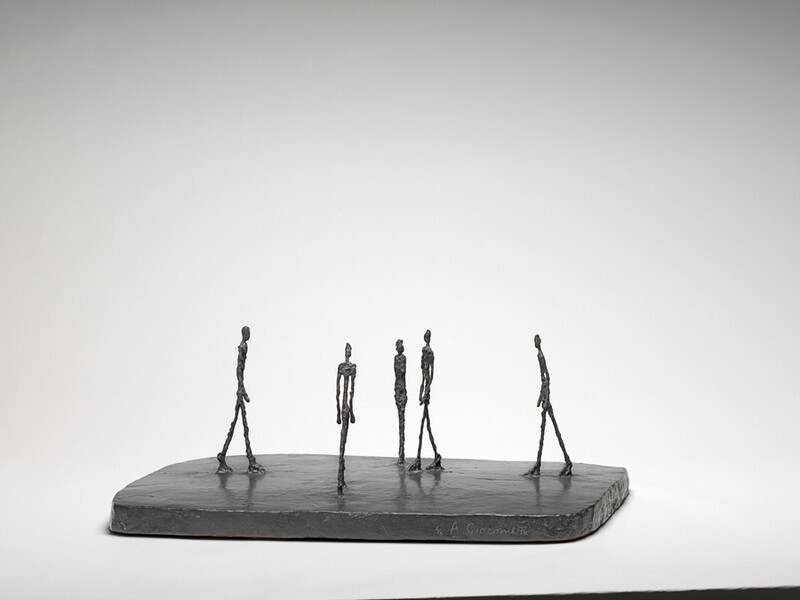 Four thin people with exaggerated, elongated legs, torsos, and arms walk across a flat, smooth, dark slab while one stands with feet together and arms by their side in this free-standing bronze sculpture. They are sparsely spaced across the base, and they all head for roughly the center of the slab. The person standing with feet together is at the center of this photograph, which shows the sculpture from one long end. The people’s bodies are textured and knobby, as if built up from small pieces of clay. The artist’s name is incised into the front face of the base to our right, and reads: “4/6 A. Giacometti.”