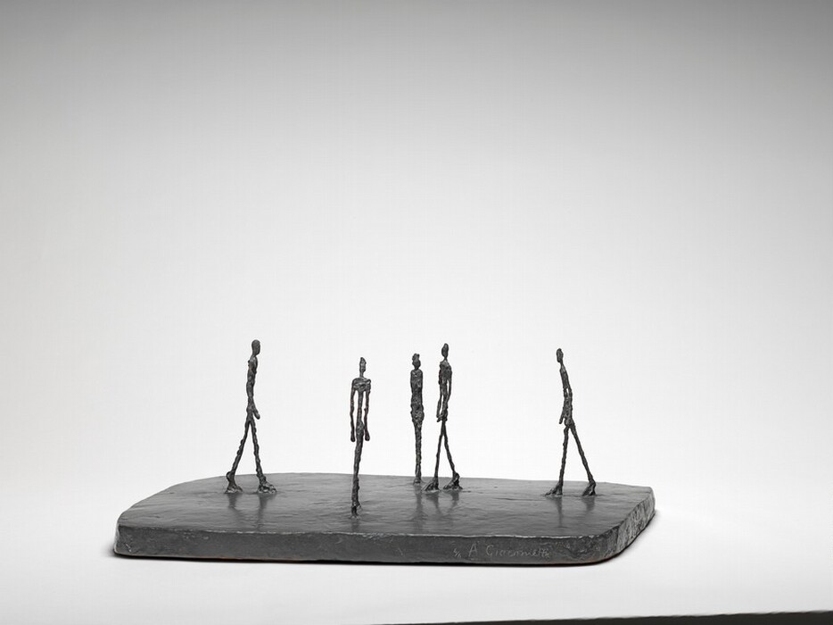 Four thin people with exaggerated, elongated legs, torsos, and arms walk across a flat, smooth, dark slab while one stands with feet together and arms by their side in this free-standing bronze sculpture. They are sparsely spaced across the base, and they all head for roughly the center of the slab. The person standing with feet together is at the center of this photograph, which shows the sculpture from one long end. The people’s bodies are textured and knobby, as if built up from small pieces of clay. The artist’s name is incised into the front face of the base to our right, and reads: “4/6 A. Giacometti.”