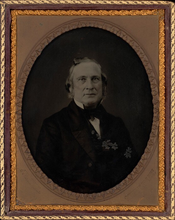 The image shows an older man seated, captured from the waist up. The man's facial features are characterized by a broad face, prominent nose, and a neutral expression. He has short, slightly wavy hair with sideburns. He is dressed in a formal dark suit, with a visible white shirt and cravat or bow tie. Marks or damage are present on the suit's shoulder area. The photograph is framed in an ornate gold case with a decorative edge. The background is plain and dark, centering the focus on the man's portrait.