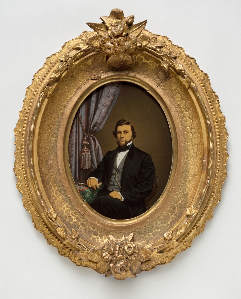 The image is a portrait of a man shown from the waist up, sitting in a formal pose. He is facing slightly to his left, with his right arm resting on a surface draped with a green fabric that has a paisley pattern. His left elbow appears to be resting on his knee. The man has a full beard and mustache, and styled hair with a side part. He is wearing a dark coat with a matching vest, white shirt, and a black bow tie. A gold chain is attached to his vest. The background features a curtain with a tassel, indicating an indoor setting. The portrait is surrounded by an oval frame with floral motifs in gold.