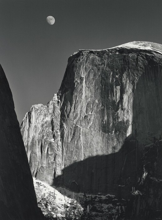 From a low vantage point, we look up at the rugged face of a tall, almost sheer rock formation with the moon above in this vertical black and white photograph. Filling most of the composition, the rock formation is slightly rounded along the top but dips down to a jagged ridge to our left. Strong light from the upper right creates deep shadows across its iron-gray surface, which is textured with long vertical streaks of white and lighter gray. An almost-full moon hovers in the otherwise clear, charcoal-gray sky above it, near the top of the image to our left of center. Formations outside our view cast deep shadows along the right edge of the photograph. A smattering of white across from us, low in the image, suggest snow on the rock there. A dark mass close to us is an ink-black silhouette rising from the lower left corner and angling off the left edge, near the upper left corner.