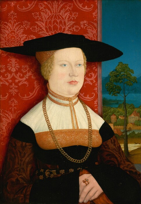 Shown from the waist up, a light-skinned woman sits angled slightly to our right in this vertical portrait painting. The woman has blue eyes, faint brows, and a sloping nose ending in a small bulb. Her upper lip is thin and her lower lip full over a slight double chin. Her blond hair is pulled back under a gold, cap-like headdress and a black, wide-brimmed hat with a shallow crown. She wears a high-necked white, black, and gold dress with intricate detailing, a gold chain necklace that falls to her waist, and a black belt with a gold clasp and medallions. She wears two gold rings set with gems on the pointer finger of her left hand, which is folded across her belly over the other hand. A scarlet-red wallcovering with pink floral designs fills two-thirds of the space behind her. The rightmost third of the background opens onto a landscape with a tall, spindly tree, a leaping animal, presumably a dog, and a bridge across a body of water leading to a town. Across another body of water, perhaps a lake, mountains are marine blue in the distance under a clear, celestial-blue sky.