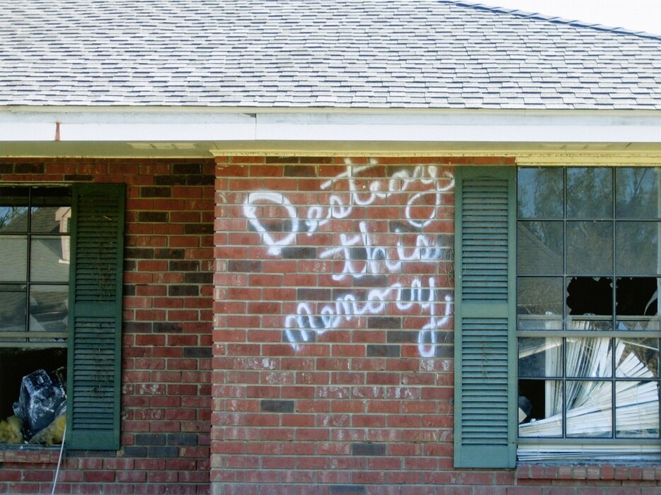 This is a photograph of a red brick wall with graffiti and broken windows. The image shows part of a building's facade featuring two windows, one on either side, that are bordered by green shutters. The wall is made of red bricks and has graffiti in white spray paint with the words "Destroy this memory" written across it. The windows have broken glass panes, with one window showing venetian blinds that are in disarray. The building's roof is visible at the top of the image, covered with grey shingles.