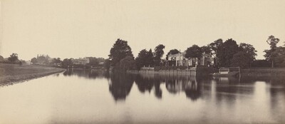 The image shows a riverside view with an estate surrounded by greenery on the other side. The horizon is high, emphasizing the reflective water surface. Trees and a house are visible on the horizon. The water reflects the surroundings. The color palette is sepia, highlighting textures and shapes in the scene.