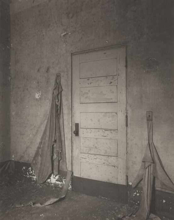 The image shows a closed, weathered wooden door with peeling paint, accompanied by two pieces of draped and tattered fabric attached to the adjacent walls. The color palette consists mainly of shades of gray, giving the image a monochromatic and aged appearance. The door is centered in the image, with one piece of fabric hanging from the wall beside the door frame and resting on the floor, while the other fabric is draped over a hook, descending toward the floor.
