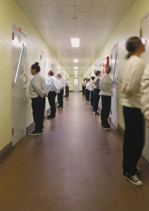 This is a photograph of a long hallway lined with doors. The hallway is lit with fluorescent ceiling lights and has a brown floor with light green walls. Each side of the hallway features several white doors with narrow vertical windows. The perspective focuses down the corridor, creating a sense of depth as it recedes to a lighted exit at the far end. The photograph captures an orderly and symmetrical architectural design.