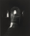 This is a photograph of light filtering through a small arched window into a dimly lit, ancient stone interior. The image captures a narrow beam of light streaming through the window, creating a soft, illuminated streak that falls upon the textured surface of the floor and wall. The room appears to be minimally lit, with the light highlighting the stonework. The play of light and shadow creates an ethereal quality, imparting a sense of quiet and stillness in this enclosed space. The overall composition emphasizes the contrast between darkness and light.