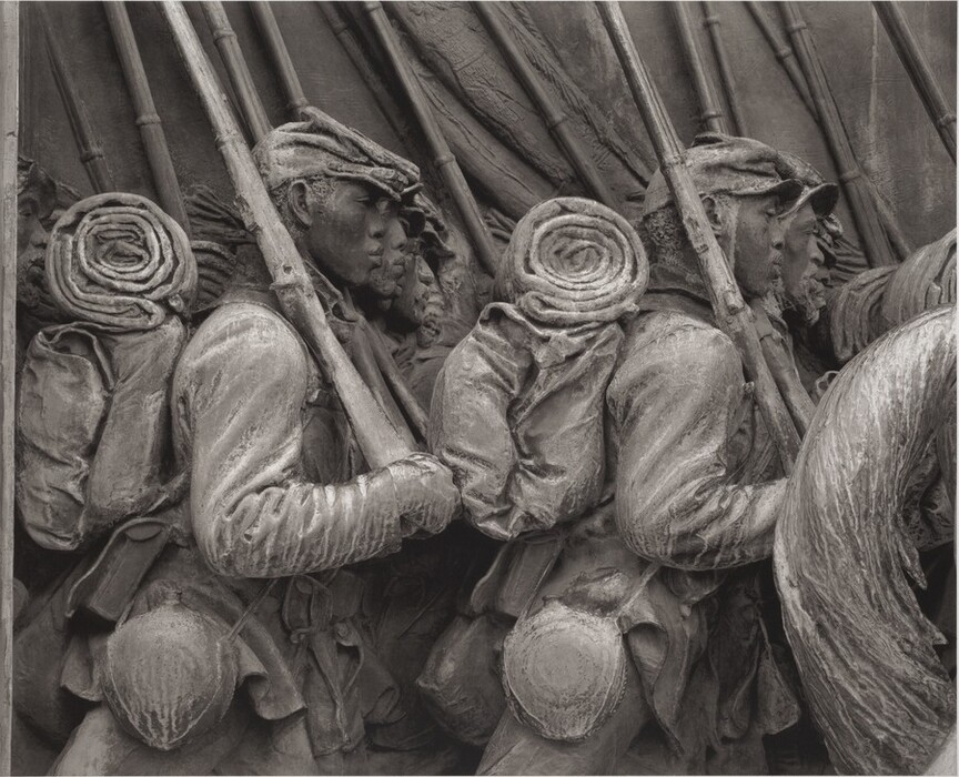 The sculpture depicts a group of soldiers marching forward while carrying rifles and wearing historical military attire. They wear caps, coats, and backpacks with rolled blankets or mats attached. All soldiers face the same direction. The sculpture is made of dark bronze with a patina for an aged look. It is sculpted in relief, showing intricate details of the soldiers' uniforms, expressions, and gear texture.