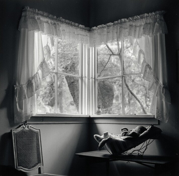 The image shows a corner window with lace-trimmed curtains, natural light entering the room. Outside, a tree is visible. Inside, a woven back chair and a wooden table with a bassinet are in view.