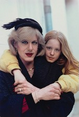 The image depicts two individuals. One adult is in the foreground, slightly turned towards the camera. There is a child next to the adult, with their arms around the adult's shoulders. The adult has light styled hair, wearing a netted fascinator hat with a feather. They have bold makeup with red lipstick and eyeliner. The child has long, straight light reddish-blonde hair and freckles. The adult is dressed in a dark coat with a hint of red underneath and accessorized with gold jewelry. The child is wearing a yellow jacket. The background is softly blurred with a light-colored wall.