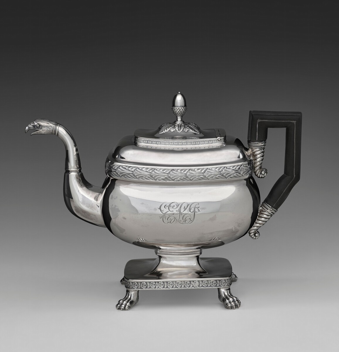 The image shows a large silver teapot with a unique spout shaped like an eagle's head, a dark handle, and intricate engravings. It has claw-footed legs, a finial on the lid, and an oval emblem on its side. The color scheme is mainly metallic silver with dark accents.