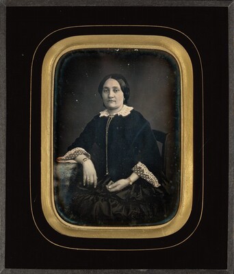 The image depicts a woman from the waist upward seated in a portrait style, facing slightly to the right. She has dark hair and is wearing a dark dress with lace collar and cuffs, accessorized with a brooch, lace detailing on the sleeves, and bracelets. The background is a dark hue, typical of daguerreotype portraits, and the photograph is framed with a gold-colored border.