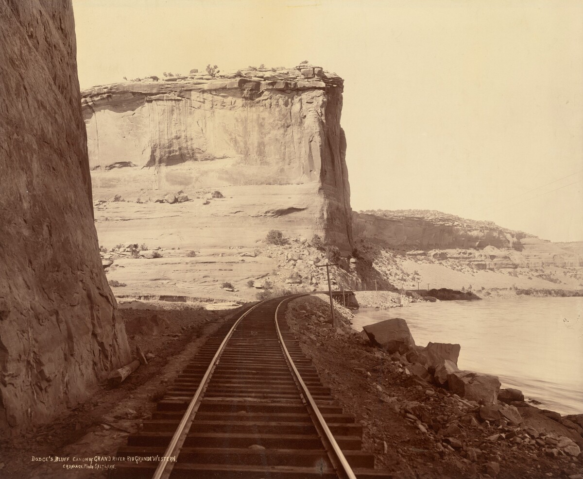 The image shows a scene of railway tracks curving to the right beside a river. The tracks are flanked by rocky terrain, with a large rock face on the left. In the distance, rugged cliffs and rock formations are visible against a clear sky. The perspective is slightly angled, giving a sense of standing at the edge of the tracks. The color palette is composed of sepia tones, creating a warm and nostalgic atmosphere. Light and shadow highlight the texture of the rocks. The image has a classical aesthetic.