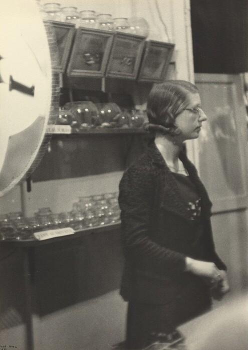 The image depicts a woman in profile view from the waist up. She is upright, facing to the right of the frame, wearing glasses, with her hair styled and pulled back. She is dressed in dark-colored clothing with a textured jacket over a top and a faintly visible necklace. She is holding what seems to be a pair of gloves in her right hand. The setting is indoors with shelves holding glass jars and square containers in the background. A large, round object with a partially visible number is in the foreground on the left side of the image.