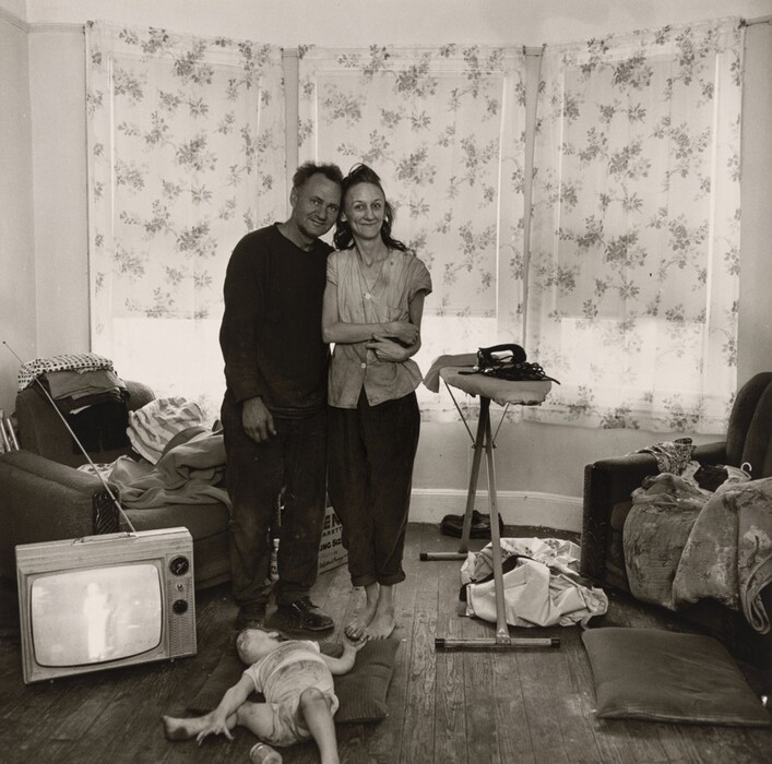 The image shows a man and a woman standing close together in the center of a room. The man is positioned slightly to the left, with his arm wrapped around the woman, who stands on his right with her arms folded. The man has unkempt hair while the woman has styled back long hair. Both wear casual, somewhat worn clothing. The man is in a dark shirt and pants, and the woman is in a light-colored, short-sleeve top with dark pants. The floor around them is wooden, and on their left side, a young child lies on the floor wearing a shirt and shorts. A television with a wire antenna is on the floor near the child, displaying an indistinct figure. Piles of clothing cover an armchair, an ironing board holds an iron and other items, and more fabric is strewn about the room. The background features a bay window with light floral curtains, allowing some light into the room, casting gentle shadows. Other assorted furniture and clutter are seen in the space.