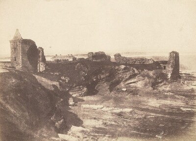 The image shows a rugged coastal landscape with a stone structure resembling a tower or fortification on the left. The stone appears weathered and aged. Rocky formations and ruins stretch along the coastline to the right, leading to the sea on the horizon. The sea forms a wide separation between the land and the sky. The image is in a sepia tone, giving it an aged appearance. The scene is barren and detailed, with warm brown and cream colors emphasizing its historical feel.