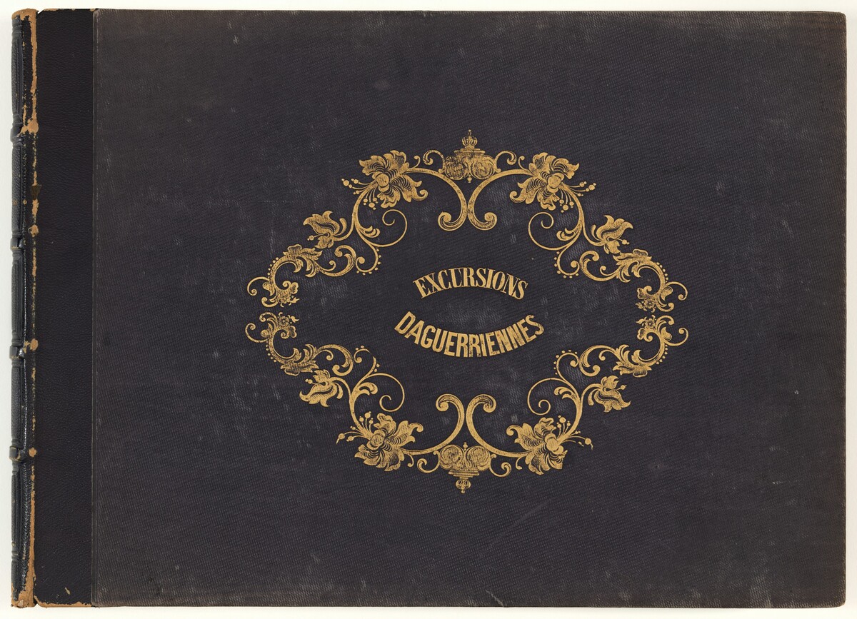 This is a photograph of a book cover. The cover is dark with elaborate gold decorations. A central ornate gold design forms an oval frame around the words "EXCURSIONS DAGUERRIENNES." The gold embellishments include scrolling floral motifs, curving filigree patterns, and a central crown-like emblem at the top of the oval. The left side of the binding shows signs of wear.
