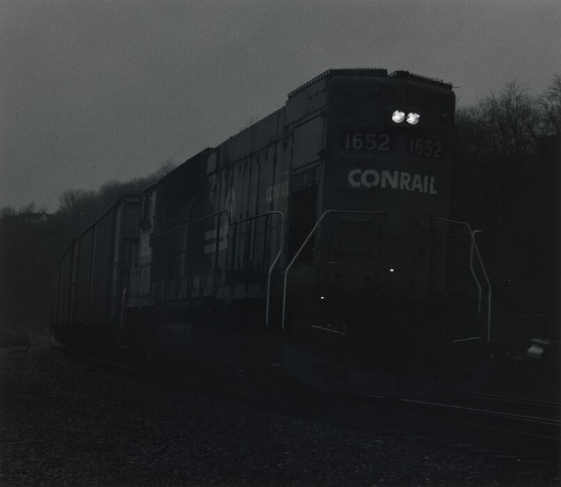 This is a photograph of a train locomotive, identified as "Conrail," emerging from a curve on a track. The image shows the front of the train displaying the numbers "1652" in text and on the lit number boards. The scene appears to be set in low light conditions. The train is a dark silhouette, and the track leads into a wooded area with barely visible barren trees lining the sides.