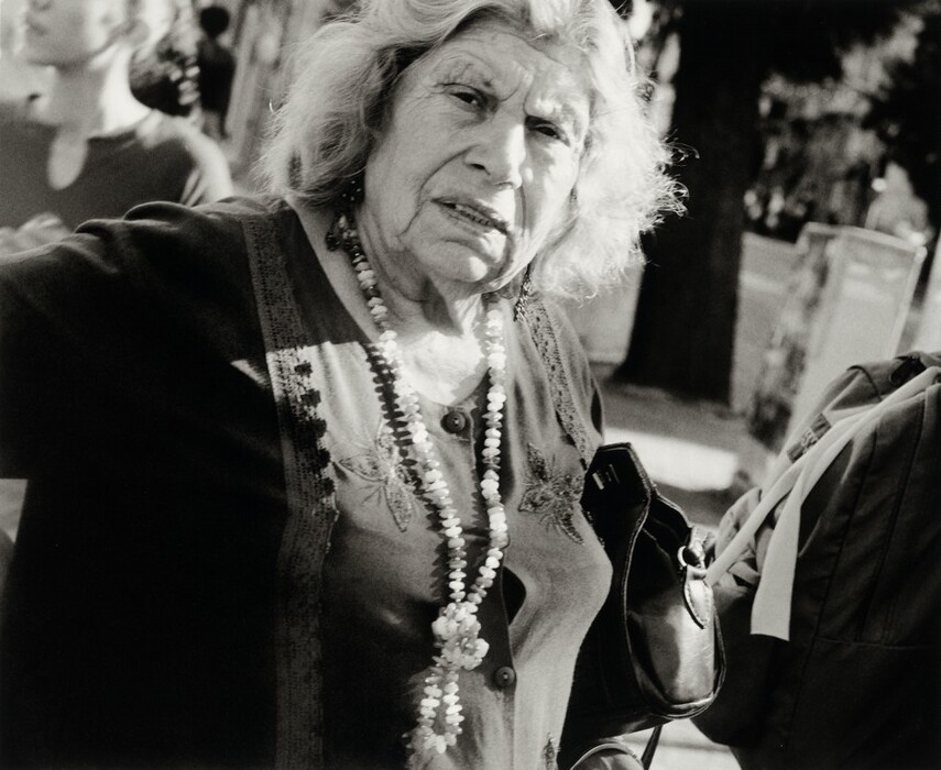 The image shows an elderly woman from the waist up, looking directly into the camera. She has shoulder-length, wavy hair, prominent cheekbones, a slightly furrowed brow, and a set expression. She wears a dark top with a patterned neckline and two large necklaces made of beads. She holds a dark handbag in her arm. The background is an outdoor scene with indistinct shapes of people and trees, suggesting a street or park environment with blurred details.
