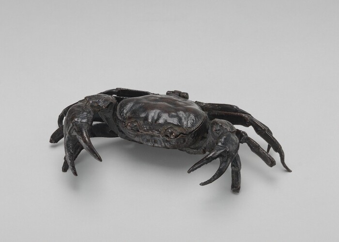 The sculpture depicts a crab with its pincers and legs extended as if in motion. It is made from a dark, metallic-looking material with a shiny, polished surface. The coloration ranges from dark brown to black and reflects light subtly across its carapace and limbs. The sculpture is detailed, mimicking the natural appearance of a crab with an intricate level of detail and realistic form. The crab's stance and intricacy suggest careful attention to anatomical accuracy.