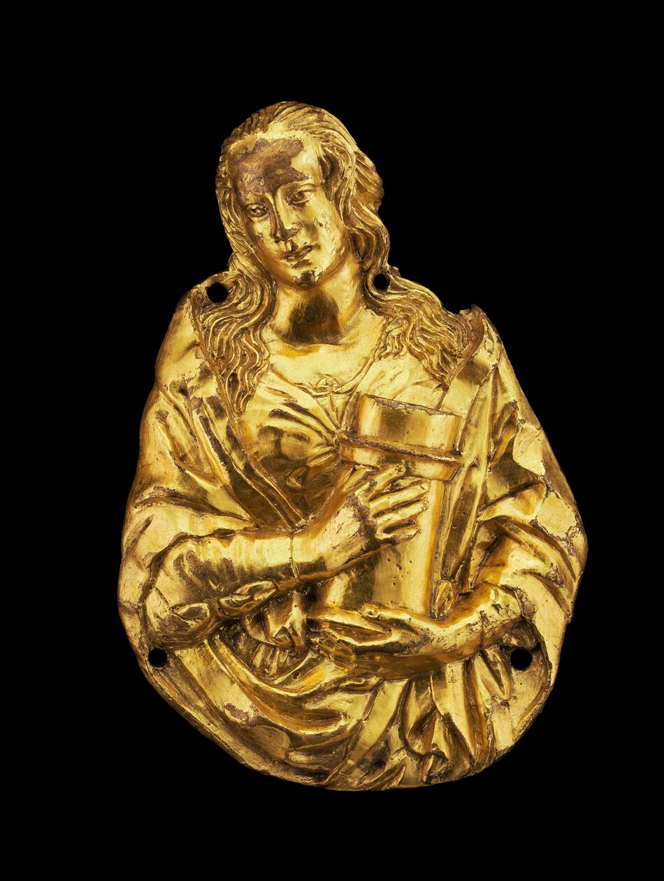 The sculpture depicts a figure with long, detailed, wavy hair cascading down to the shoulders. The figure appears to be wearing a flowing garment with draped sleeves. In the figure's hands is a cylindrical object. Made from a metallic material, the sculpture is gilded with a bright gold color. The surface has a polished finish, reflecting light. The sculpture is relatively flat.