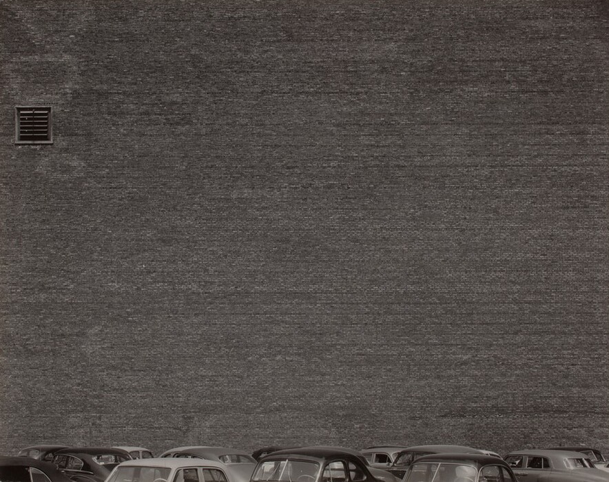 The image shows a large brick wall filling most of the frame, viewed from a slightly elevated angle. The horizon is low, with parked cars at the bottom. The wall dominates the background with a uniform pattern of small bricks. The tops of cars are visible at the bottom of the image. A small square vent breaks the wall's surface in the upper left. The image is in black and white, highlighting the scene's starkness and simplicity with emphasis on geometric shapes and patterns.