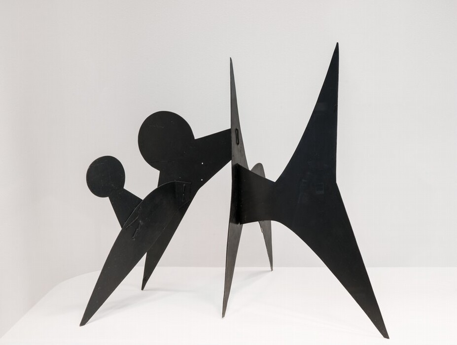 The sculpture features abstract forms combining circular and triangular shapes in a dynamic arrangement. It is made from black metal, giving it a sleek appearance. The sculpture consists of sharp angles and pointed edges, standing on three legs, with two circular elements protruding from one side. It has an asymmetrical form and appears to balance delicately despite its seemingly unstable shape, creating an engaging and visually interesting piece against a white background.