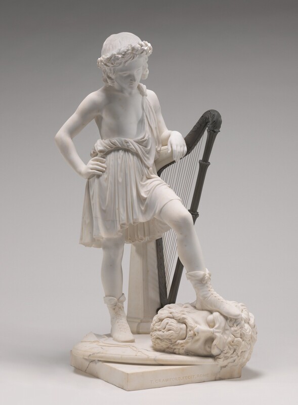 A smooth-skinned boy, David, looks down at a severed head lying at his feet in this free-standing white marble sculpture. David’s body and head are angled slightly to our right in this photograph, and we look directly onto the large face of the severed head, which belonged to Goliath. David’s head is bowed, and he wears a ring of flowers over shoulder-length curls. His right hand, to our left, rests on that hip, and his other elbow is propped on a chest-high, bronze harp. Cloth drapes over David’s far shoulder, though his chest is otherwise bare. The fabric twists around the waist and falls to his knees. High boots are laced up over the ankles. One foot rests squarely on the temple of Goliath’s severed head. Goliath’s heavy brows are deeply furrowed over closed, deep-set eyes. His cheeks are lined, his mouth downturned in a bushy, curly beard. A slingshot lies on the base next to Goliath’s head. They are on a thin, disk-like base atop a thicker square base, all carved from marble. The artist inscribed the front face of the lower base to read, “T. CRAWFORD. FECIT. ROME. 1848.”