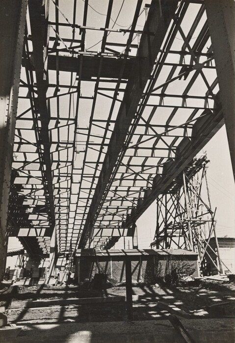 In this vertical grayscale photograph, the angular steel framework of a large industrial structure stretches across the scene. The structure is composed of intersecting beams and trusses, seen from below, creating a pattern of lines and shapes against the sky. Shadows of the framework are cast on the ground below. On the right side, additional scaffolding supports the structure, with low buildings partially visible in the background.