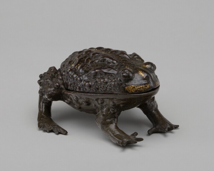 The sculpture depicts a frog in a crouched position. The frog is crafted from a dark material, possibly bronze or a similar metal, which gives it a deep brown, almost black appearance with some glossy surfaces. There are detailed textures across its back, mimicking the bumpy skin of a real frog. There are also some subtle touches of gold glinting across its forehead and around its eyes. This sculpture resembles a life-sized representation of a frog. Its features such as the legs, eyes, and textured skin give it a naturalistic presence.