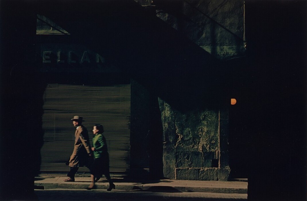 This is a photograph of an urban street scene captured in low light. The image shows a street with a closed shop front on the left, its metal rolling shutter casting stripes of shadow on the pavement. To the right is a textured wall with dark shadows where ambient light struggles to reach. Above, a small section of an angular metal structure contributes to the industrial atmosphere of the scene. Patches of light suggest a partly obscured sign reading "ELLAN" on the left, while an orange sphere adds a splash of warm color to the right. The play of light and shadow creates a mysterious and atmospheric narrative.