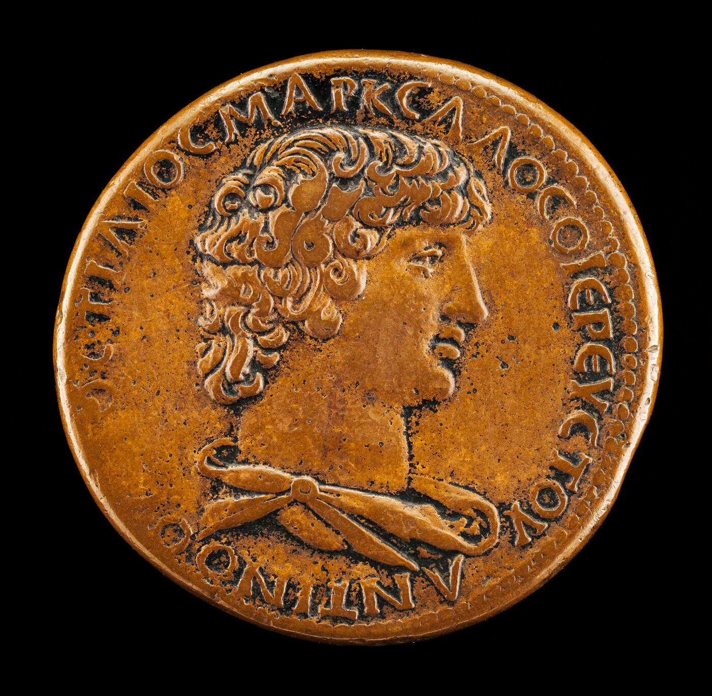 The sculpture is a profile portrait of a man with curly hair, depicted in relief. His hair is styled, and he is wearing a draped garment fastened with a brooch. It is set on a round coin with raised letters encircling the portrait. The sculpture has a golden-brown color, suggesting it is made of bronze or a similar metal. The details are well-defined, with shadows accentuating the man's features. The overall appearance is that of a classical or ancient coin, with artistic and inscriptional elements conveying historical or cultural significance.