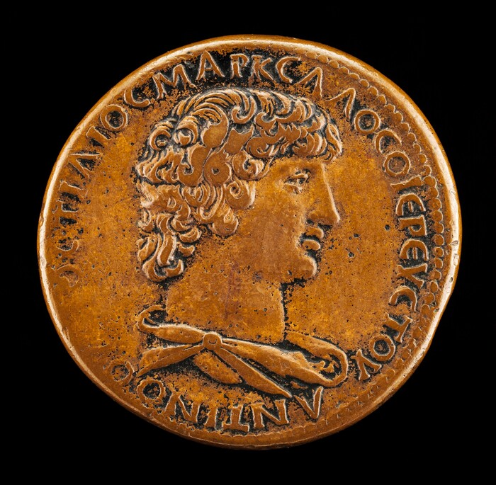 The sculpture is a profile portrait of a man with curly hair, depicted in relief. His hair is styled, and he is wearing a draped garment fastened with a brooch. It is set on a round coin with raised letters encircling the portrait. The sculpture has a golden-brown color, suggesting it is made of bronze or a similar metal. The details are well-defined, with shadows accentuating the man's features. The overall appearance is that of a classical or ancient coin, with artistic and inscriptional elements conveying historical or cultural significance.