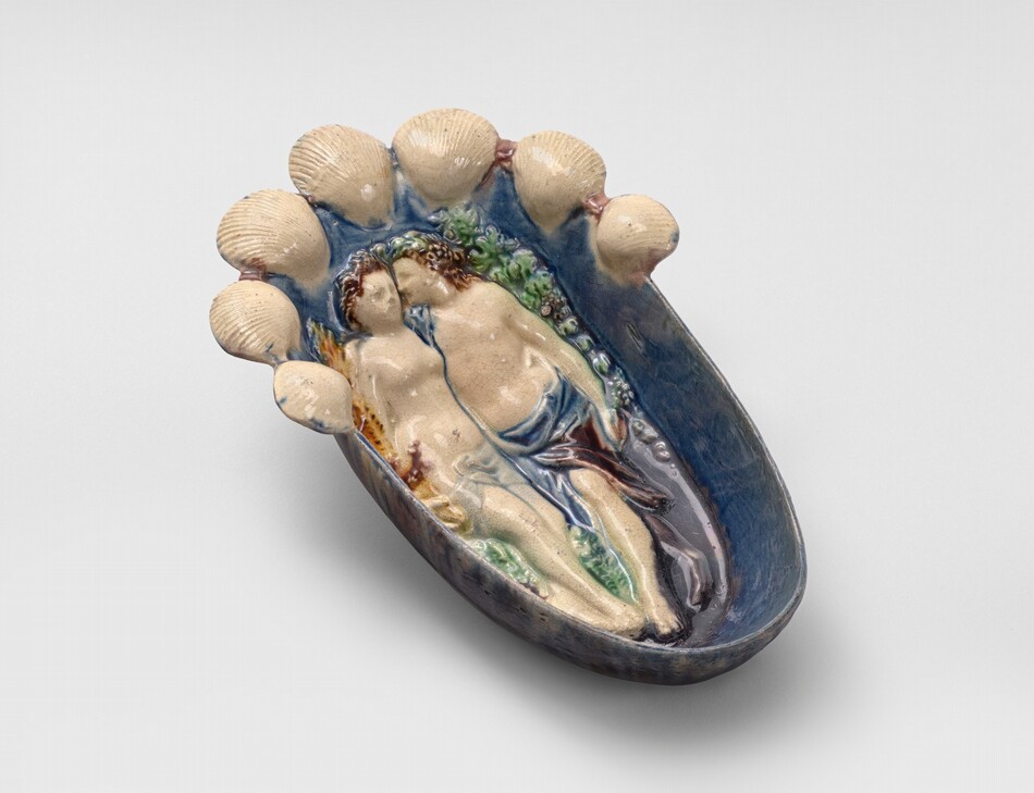 The sculpture is a shallow dish with two reclining figures at its center. The figures are human and intertwined, surrounded by small white seashell-shaped elements. The dish is made of ceramic with various colors. The material has a three-dimensional appearance and is detailed. The colors used are greens, blues, and flesh tones with a deep blue shade on the outer rim, giving it a lush and colorful look with naturalistic elements.