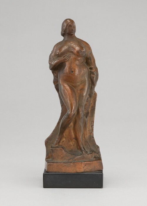 The sculpture depicts a nude female figure in a standing pose. The figure has a slightly turned stance, with one leg crossed in front of the other and an arm across her midsection, creating a classic contrapposto posture. The sculpture is crafted from a material that resembles bronze, giving it a warm, brownish-gold color with a subtle sheen. The surface of the sculpture is smooth, with gentle curves that define the figure's form. It stands on a rectangular, black base. The details in the sculpture are simplified, with a focus on capturing the overall shape and posture of the figure.