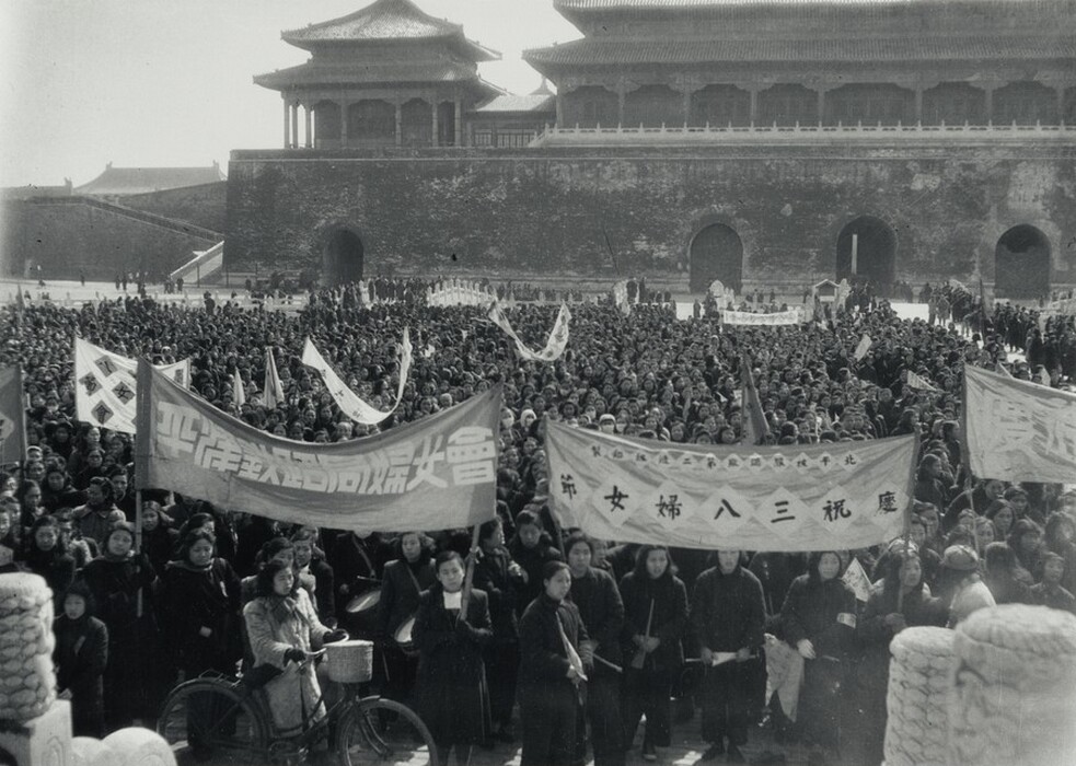 This is a photograph of a large public demonstration in front of a historical architectural structure. The image depicts a vast crowd gathered in an open area adorned with banners and flags featuring inscriptions in an East Asian script. The structure in the background bears features typical of traditional East Asian architecture, with tiered roofing and large gates. The atmosphere suggests a significant gathering, as people fill the space holding signs, while one person is notably standing with a bicycle at the front. The masses appear to be organized and engaged, focused in a common direction. The photograph captures the crowd's density and the backdrop's grandeur, offering a snapshot of a historical moment.