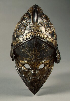 This is an ornate helmet made of dark metal with gold-colored details. It features intricate designs and patterns throughout, including symmetrical swirling and floral designs, as well as designs on the faceguard that look similar to an animal’s face, including a snarling mouth and eye holes. The lighting emphasizes the embossed and engraved elements of the helmet.