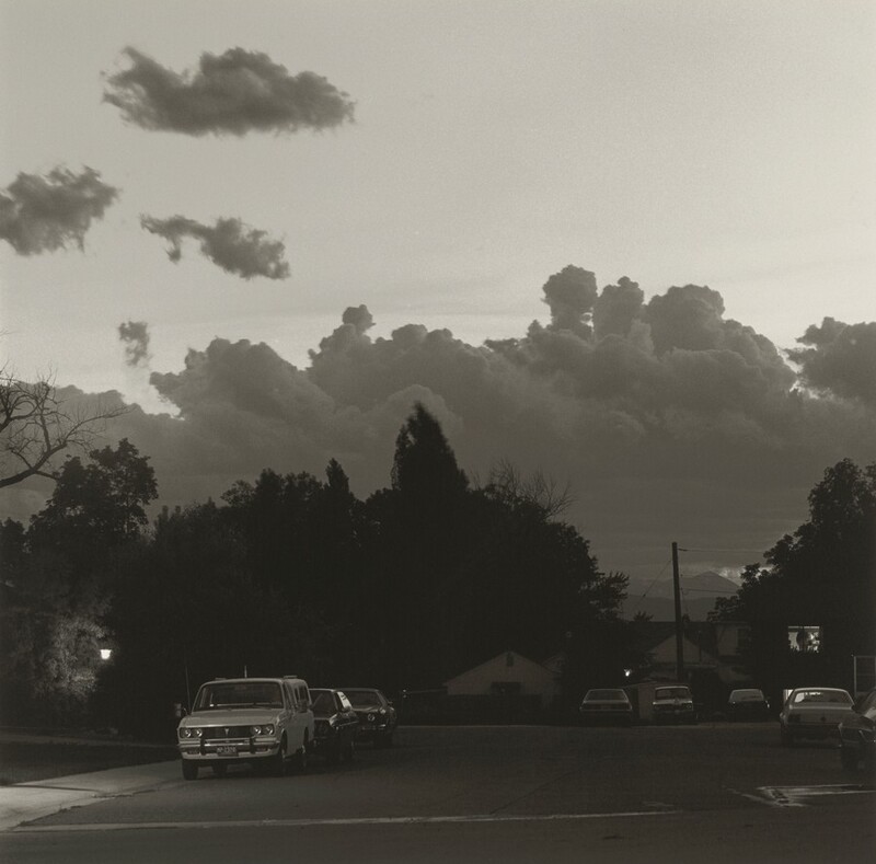 A dense bank of clouds kicks up over a tree-lined neighborhood street in this square black and white photograph. We are on the street with cars are parked to our left and right. A few houses are tucked in among the dark trees across from us. The band of clouds takes up the middle third of the picture above the street. A few puffs float near the top left corner of the photograph in the otherwise empty sky.