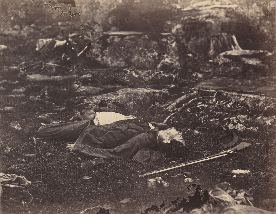 This is a black-and-white photograph of a war-torn landscape with a fallen figure on the ground. The scene is characterized by rough terrain of rocks and muddy, uneven earth. In the center of the image, a figure is lying flat among the grass and dirt, with a gun laying on the ground next to them. They have light skin, dark hair, and a dark jacket open to show a white shirt. There are many shapes in the background that could be other bodies or the remnants of clothing or belongings.