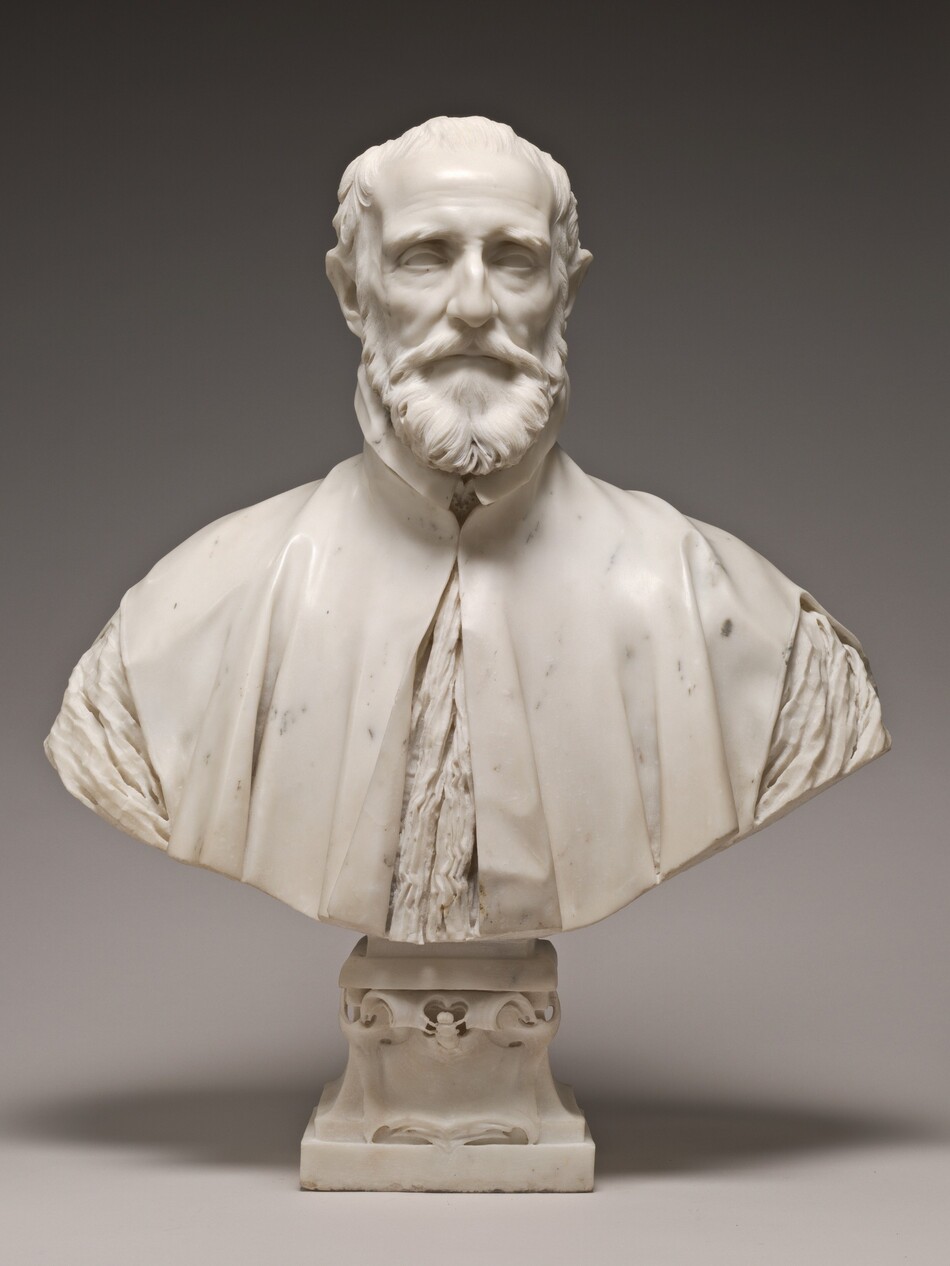 This free-standing, white marble sculpture shows the head, shoulders, and upper chest of a balding, bearded man. In this photograph, his body faces us and he looks straight ahead. Bushy eyebrows arch low over deeply set eyes, and his forehead hollows inward at his temples. He has high cheekbones and the skin of his cheeks draws slightly in. His thin lips are set in a straight line within a full beard. His high-necked robe falls slightly open over his chest and is cut also away over the shoulders to show the crinkled fabric of the garment beneath. The bottom edge of the sculpture curves upward in a very shallow U across the bottom, and is supported by a square pedestal foot decorated with scroll designs. The background behind the bust lightens from charcoal gray along the top edge to pale gray across the bottom, and the sculpture casts a shadow behind its base.