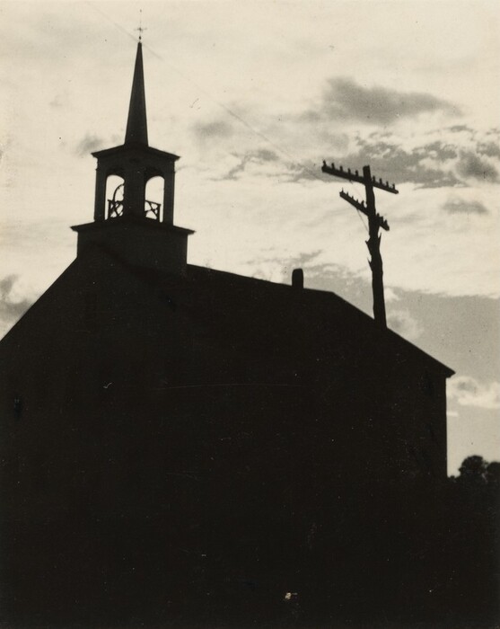 The image shows a view from ground level looking up at a silhouetted building with a steeple. The building occupies most of the lower portion of the image, with a telephone pole slightly to the right. The sky filled with scattered clouds provides a backdrop to the dark shapes. The image has a stark contrast between the dark building and the lighter sky, emphasizing the outlines of the architectural features and the telephone pole. The color palette is monochrome with various shades of black, white, and gray, creating a dramatic and composed visual impact.