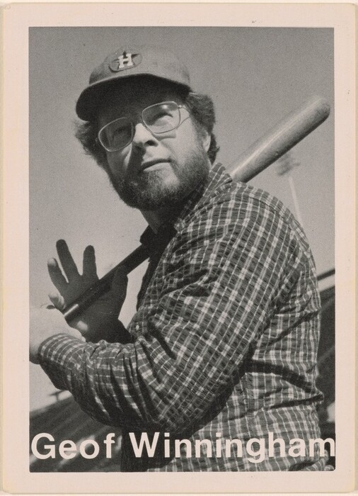 The image shows a man's upper body from the waist up. He is positioned with his left shoulder angled towards the camera, holding a baseball bat over his right shoulder. He has a full beard and mustache and wears glasses, providing a clear view of his facial features. The man is wearing a checkered shirt and a cap with a logo. The background is a clear sky with what seems to be the top of a baseball stadium light structure barely visible in the corner.