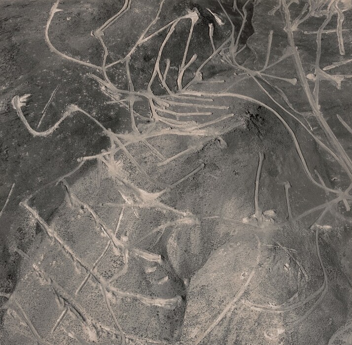 This is a photograph of a desert landscape with a series of spiral and linear pathways. The image shows an aerial view of the terrain, with intricate lines etched into the soil. The pathways traverse through varied terrain, some sparsely vegetated with signs of flat arid desert, and others showing slightly elevated landforms. The predominant color is a muted, earthy tone, suggesting a dusty environment typical of arid regions. The pathways form complex, interconnected shapes, adding elegance to the natural landscape.