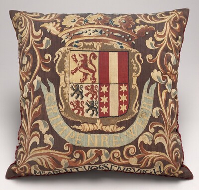 This is a photograph of a decorative embroidered cushion. The cushion features a detailed coat of arms in the center, showcasing a lion, stars, and various stripes and patterns within an ornate shield. The crest is surrounded by floral and scroll motifs in colors like gold, red, blue, and brown. The design includes a banner below the shield with text. The craftsmanship is intricate, highlighting woven patterns and textures.
