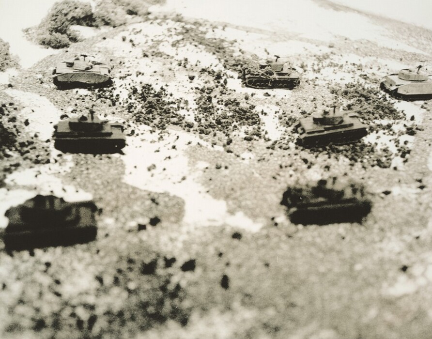 This is a photograph of model tanks on a miniature landscape. Six model tanks are placed on a textured terrain that appears rugged, possibly representing a battlefield. The ground is covered with small, pebble-like material resembling dirt and vegetation. The tanks are positioned at different angles, some facing the camera and others facing away. The image is in black and white, giving a vintage feel. The composition and focus on the miniature scale create depth and perspective.