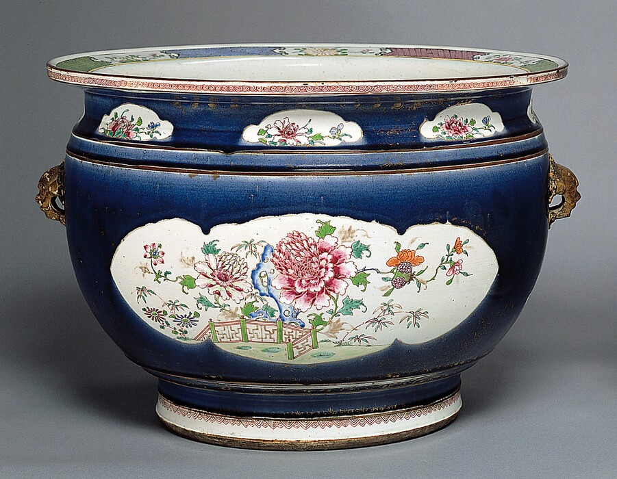 The sculpture is an ornate, decorative vessel resembling a large ceramic bowl or pot. Its surface features intricate floral designs painted in shades of pink, red, and orange, with hints of green leaves and a small brown fence around the flowers. The main body of the vessel is a deep cobalt blue color and has a smooth, shiny surface. The vessel has small golden-toned handles on each side. The setting is neutral, with a plain grayish backdrop.