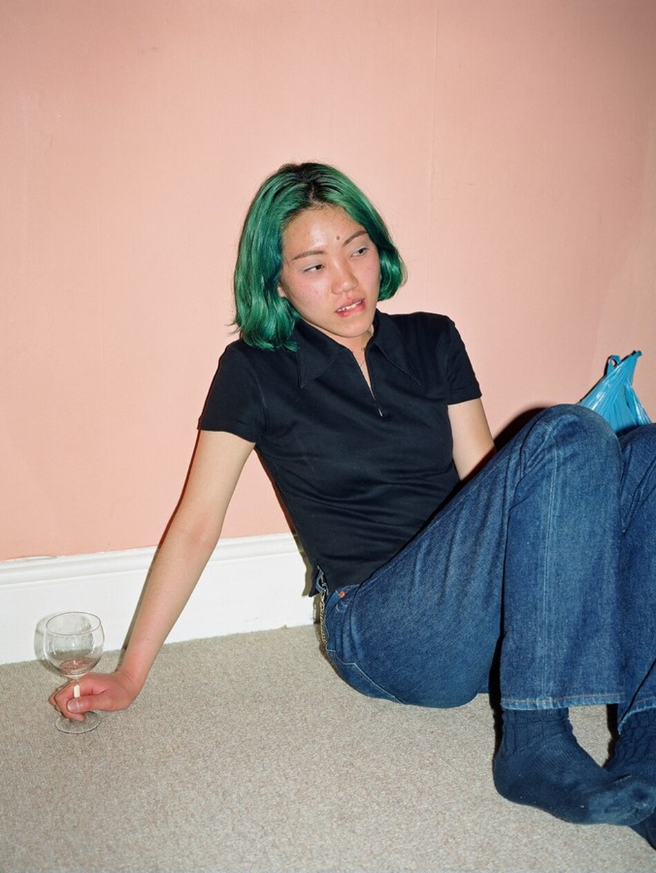 This photograph shows a person sitting on the floor with their body leaning against a peach-colored wall. Their head is tilted, and a portion of their torso and legs is visible. The person has chin-length green hair with dark roots. They are wearing a short-sleeve, dark-colored collared shirt and blue jeans, with dark socks visible. The person is holding an empty wine glass in one hand, which is placed on the light-colored carpet. A blue plastic bag is beside them. The background features a plain peach wall with a white baseboard.