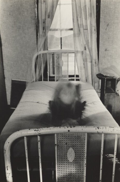 This is an image of a metal-framed bed in a sparsely decorated room. A window with thin curtains is at the head of the bed. The bed has a simple cotton sheet on it. There is a blurry object on the bed. A piece of furniture or shelf with folded clothes is on the right. Light comes in through the window, creating soft shadows. The room gives off a feeling of quietness and seclusion.